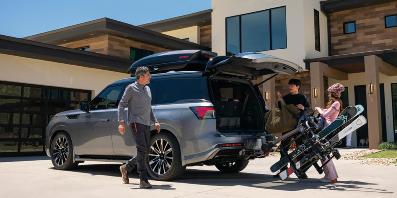 A family unloading ski gear from the back of a silver Infiniti QX80 SUV in front of a modern home.